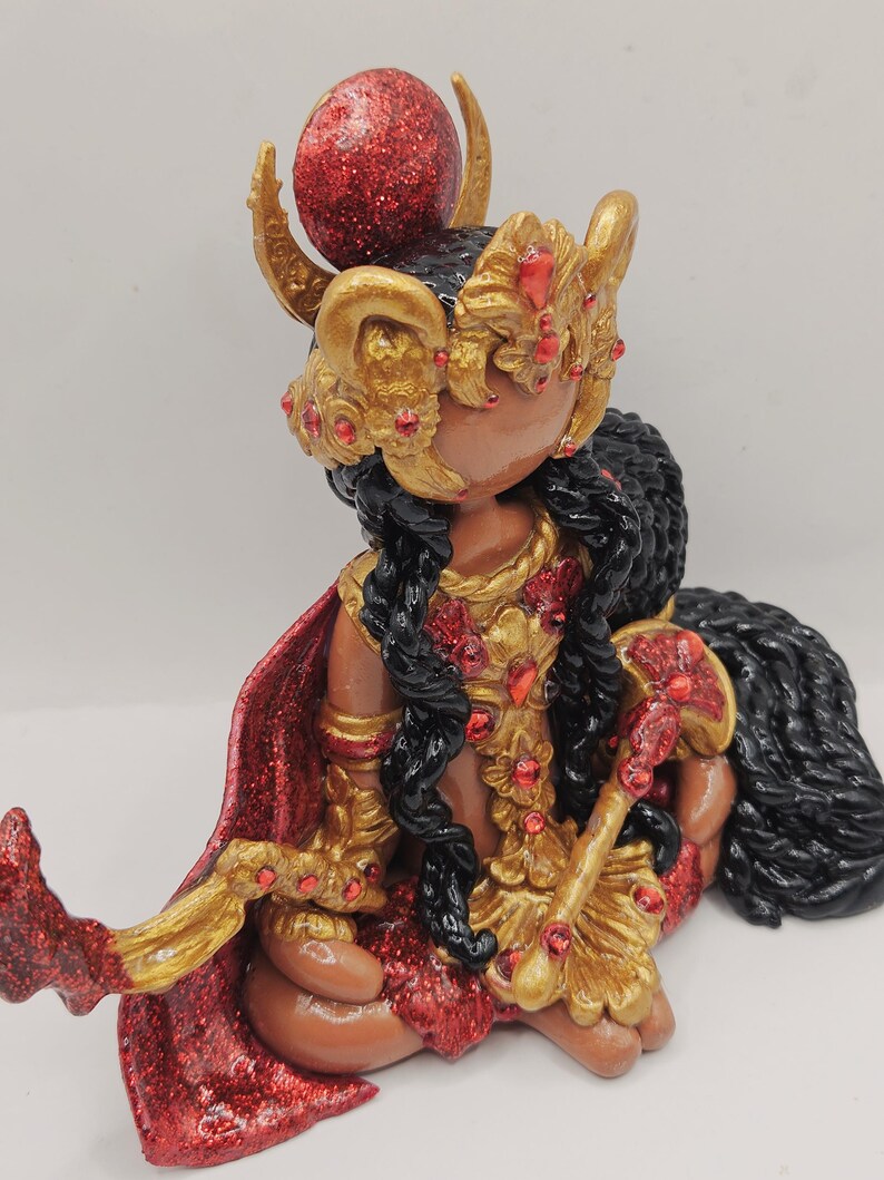 May include: A handcrafted figurine of a seated figure with dark skin, wearing a gold and red ornate headdress and clothing. The figurine has long black hair and a red cape. The figure is holding a gold staff with red accents. The figurine is on a white surface.