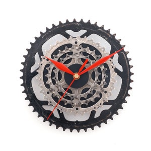 Può includere: Un orologio nero e argento con lancette rosse, realizzato con ingranaggi per biciclette. Il quadrante dell'orologio è un cerchio nero con un anello argentato attorno. Gli ingranaggi sono disposti in un motivo circolare concentrico.