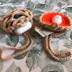 May include: Two crocheted pie-shaped pouches with a tan cord. One pouch is filled with a white AirPod case and the other pouch is orange with a white swirl on top.