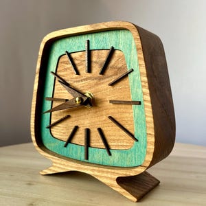 May include: A wooden clock with a teal blue face and gold hands. The clock has a square shape with rounded corners and a stand.