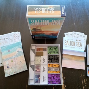 Puede incluir: Una caja blanca con un inserto de juego para el juego de mesa Salton Sea. El inserto tiene compartimentos para los componentes del juego, incluidas cartas, fichas y losetas. La caja está etiquetada con el título del juego y el nombre del editor.
