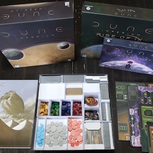 May include: A collection of board game components for the Dune: Imperium board game. The components include a game board, cards, tokens, and dice. The game is set in the universe of the Dune novels and film.