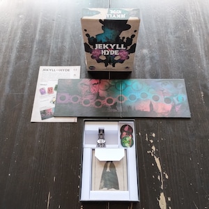 Könnte beinhalten: Eine Brettspielbox mit einem Schwarzweißbild eines Männergesichts mit einem Spritzer violetter und grüner Tinte. Die Box ist geöffnet und das Spielbrett ist sichtbar. Das Spielbrett ist schwarz mit einem violetten und grünen Design. Das Spielbrett ist in der Mitte gefaltet. Die Spielfiguren sind in der Box sichtbar. Das Spiel heißt "Jekyll & Hyde".