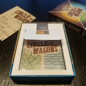 May include: A blue box with a white insert and a card game inside. The card game is called "Circle the Wagons" and features a colourful design with trees, water, and a sunset.