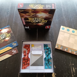 Puede incluir: Un juego de mesa llamado "Match of the Century" con un tablero de madera marrón, piezas de ajedrez rojas y azules, y una caja blanca con instrucciones y una descripción histórica.