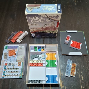Puede incluir: La caja del juego de mesa The White Castle con sus componentes, incluyendo un tablero de juego, piezas de madera y dados. La caja presenta una ilustración colorida de un castillo blanco y un paisaje japonés.