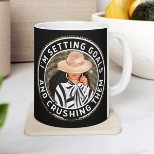 May include: White ceramic mug with a black background and gold glitter accents. The mug features a black and white illustration of a woman wearing a hat and a striped shirt. The text "I'm Setting Goals And Crushing Them" is written in a circle around the woman.