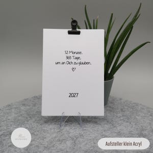Pu&ograve; includere: Una carta rettangolare bianca tenuta da una clip nera su un supporto in acrilico trasparente. La carta presenta un testo nero che recita 12 Monate. 365 Tage, um an Dich zu glauben. con un piccolo simbolo a cuore e l'anno 2027 in basso.