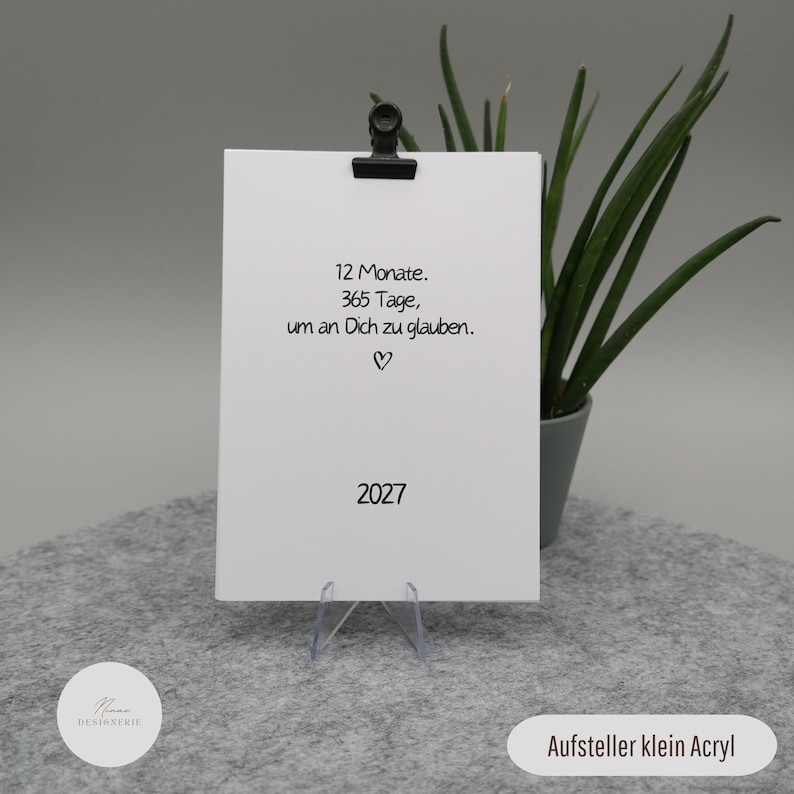 Pu&ograve; includere: Una carta rettangolare bianca tenuta da una clip nera su un supporto in acrilico trasparente. La carta presenta un testo nero che recita 12 Monate. 365 Tage, um an Dich zu glauben. con un piccolo simbolo a cuore e l'anno 2027 in basso.