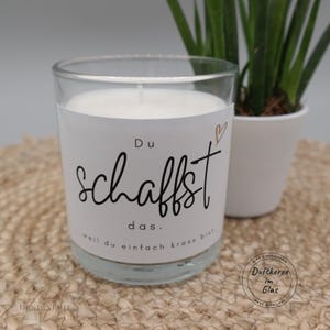 Könnte beinhalten: Eine Duftkerze im Glas mit weißem Wachs und einem weißen Etikett, das mit dem schwarzen Text Du schaffst das...weil du einfach krass bist und einem kleinen goldenen Herzsymbol bedruckt ist. Die Kerze hat einen einzelnen weißen Docht.