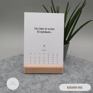 Pu&ograve; includere: Una scheda calendario rettangolare bianca con il testo in tedesco Das Leben ist zu kurz f&uuml;r irgendwann, una piccola icona a forma di cuore e il mese di gennaio con le date, tenuta in verticale da un supporto a blocco di legno chiaro.