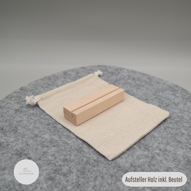 Pu&ograve; includere: Un supporto rettangolare in legno di colore chiaro con una stretta fessura orizzontale sulla superficie superiore, accompagnato da un piccolo sacchetto in tessuto beige con cordoncini bianchi, etichettato con il testo Aufsteller Holz inkl. Beutel.