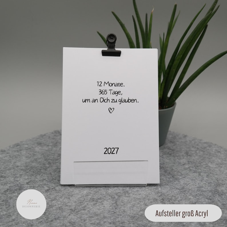 Pu&ograve; includere: Una carta rettangolare bianca tenuta da una clip nera su un supporto in acrilico trasparente. La carta presenta un testo nero che recita 12 Monate. 365 Tage, um an Dich zu glauben. con un piccolo cuore e l'anno 2027 stampato in basso.