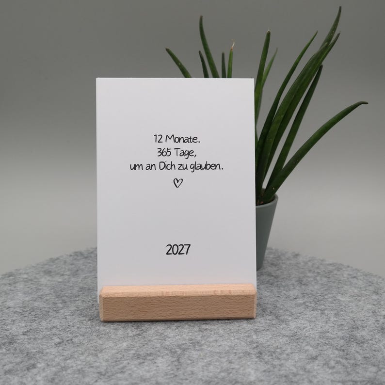 Pu&ograve; includere: Un cartoncino rettangolare bianco inserito in un supporto di legno chiaro. Il cartoncino presenta una scritta nera che recita 12 Monate. 365 Tage, um an Dich zu glauben. seguita da un piccolo simbolo a forma di cuore e l'anno 2027 in basso.