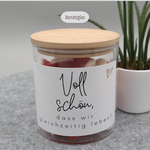 Könnte beinhalten: Ein klares Glas mit Holzdeckel, gefüllt mit roten und weißen Süßigkeiten. Ein weißes Etikett mit schwarzer Schrift lautet "Voll schön, dass wir gleichzeitig leben!". Ein kleines goldenes Herz ist sichtbar. Eine kleine Topfpflanze befindet sich im Hintergrund.