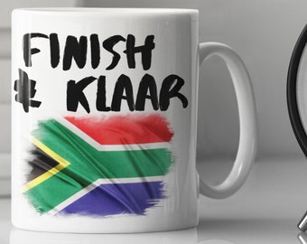 Könnte beinhalten: Eine weiße Keramiktasse mit einer schwarz-weißen Grafik, auf der "Finish Klaar" steht, und einem verblassten südafrikanischen Flaggen-Design.