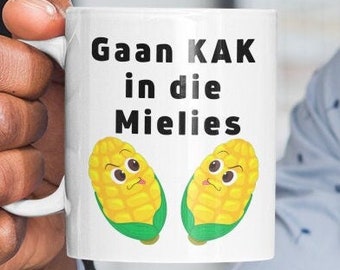 Könnte beinhalten: Eine weiße Keramiktasse mit einem schwarz-weißen Design. Die Tasse zeigt den Text "Gaan KAK in die Mielies" und zwei Comic-Maiskolben mit Gesichtern.