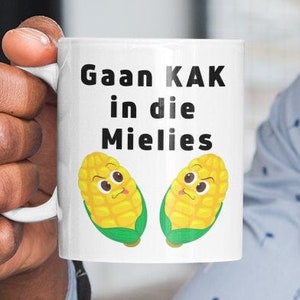 May include: White ceramic mug with a black and white design. The mug features the text "Gaan KAK in die Mielies" and two cartoon corn cobs with faces.