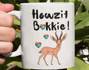 Könnte beinhalten: Weiße Keramiktasse mit einer schwarz-weißen Grafik eines Springbocks und dem Text "Howzit Bokkie!" mit drei roten Herzen, die die südafrikanische Flagge zeigen.
