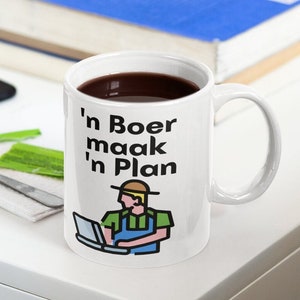 Peut inclure: Une tasse en céramique blanche avec du texte noir qui dit "'n Boer maak 'n Plan" et une illustration de dessin animé d'un agriculteur utilisant un ordinateur portable.