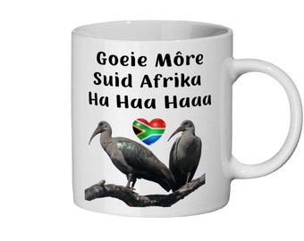 May include: White ceramic mug with a black and white graphic of two birds on a branch. The graphic also includes a heart with the South African flag. The text on the mug reads "Goeie Môre Suid Afrika Ha Haa Haaa".