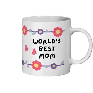 以下が含まれることがあります： 「WORLD'S BEST MOM」の文字が黒で書かれた白いセラミックマグ。ピンクの花、紫の花、ピンクの蝶で飾られています。マグには白いハンドルが付いています。