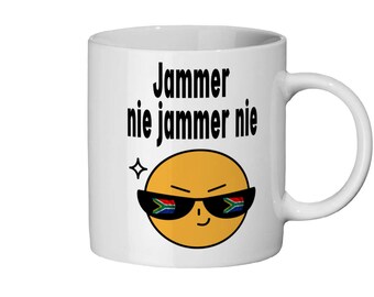 May include: White ceramic mug featuring a yellow smiley face sporting sunglasses with the South African flag design on the lenses. The text "Jammer nie jammer nie" is printed in black above the smiley face.