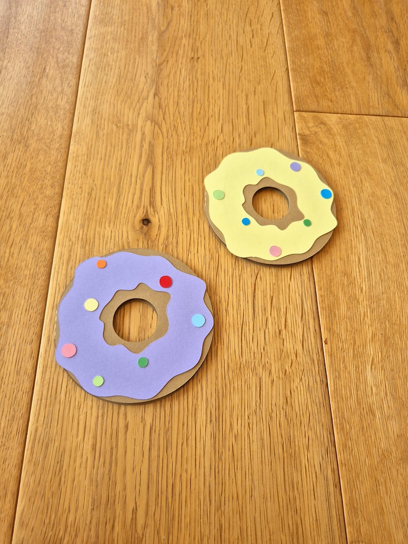 May include: Two paper donuts with colourful sprinkles. One donut is purple with a brown centre and the other is yellow with a brown centre.