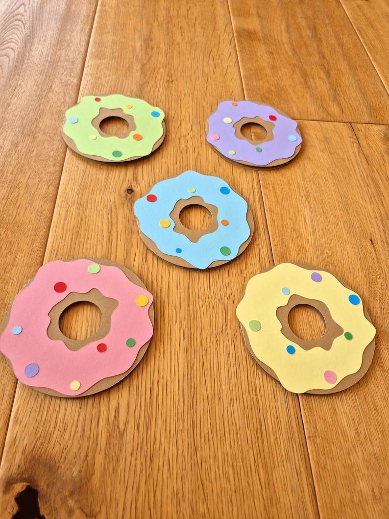 May include: Five paper donuts with different coloured frosting and sprinkles. The donuts are light green, purple, light blue, pink, and yellow.
