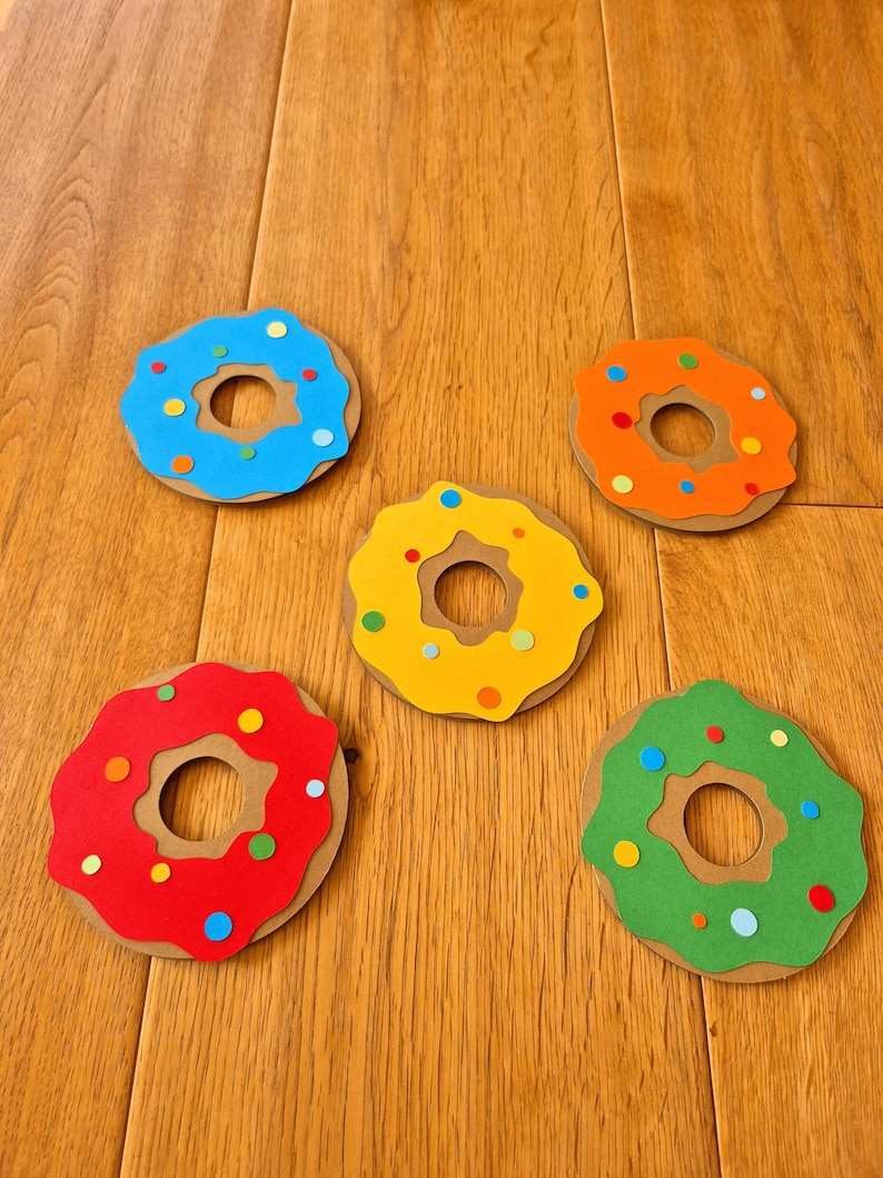 May include: Five paper donuts in different colours: blue, yellow, red, green, and orange. Each donut has a brown centre and colourful sprinkles.