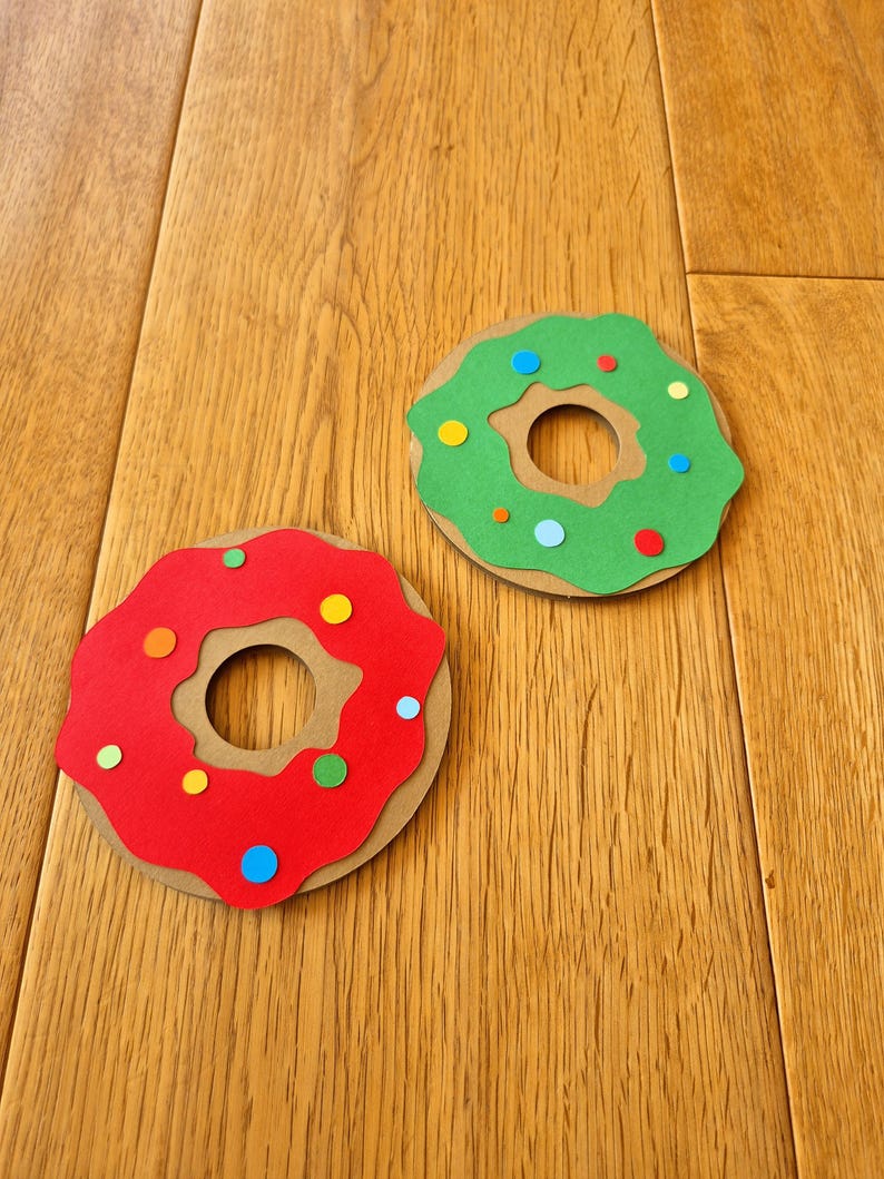 May include: Two paper donuts, one red and one green, with colourful sprinkles. The donuts are on a wooden surface.