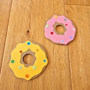 May include: Two paper donuts, one yellow with brown icing and the other pink with brown icing, decorated with colourful sprinkles.
