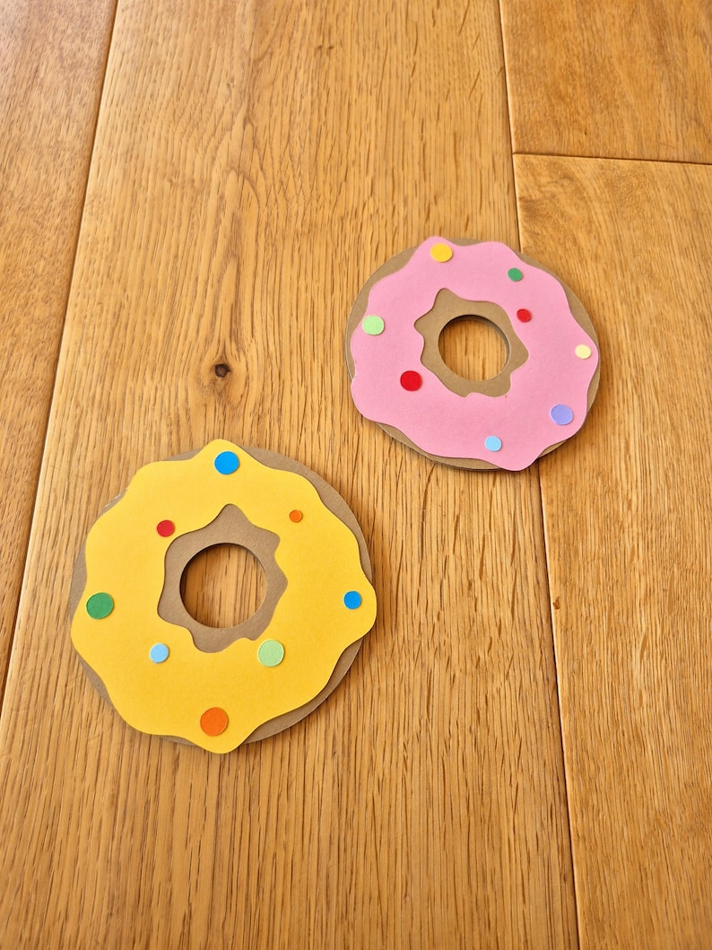 May include: Two paper donuts, one yellow with brown icing and the other pink with brown icing, decorated with colourful sprinkles.