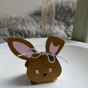 May include: A brown paper bunny-shaped box with pink ears and a gray ribbon. The box has a white face with black eyes and a nose.