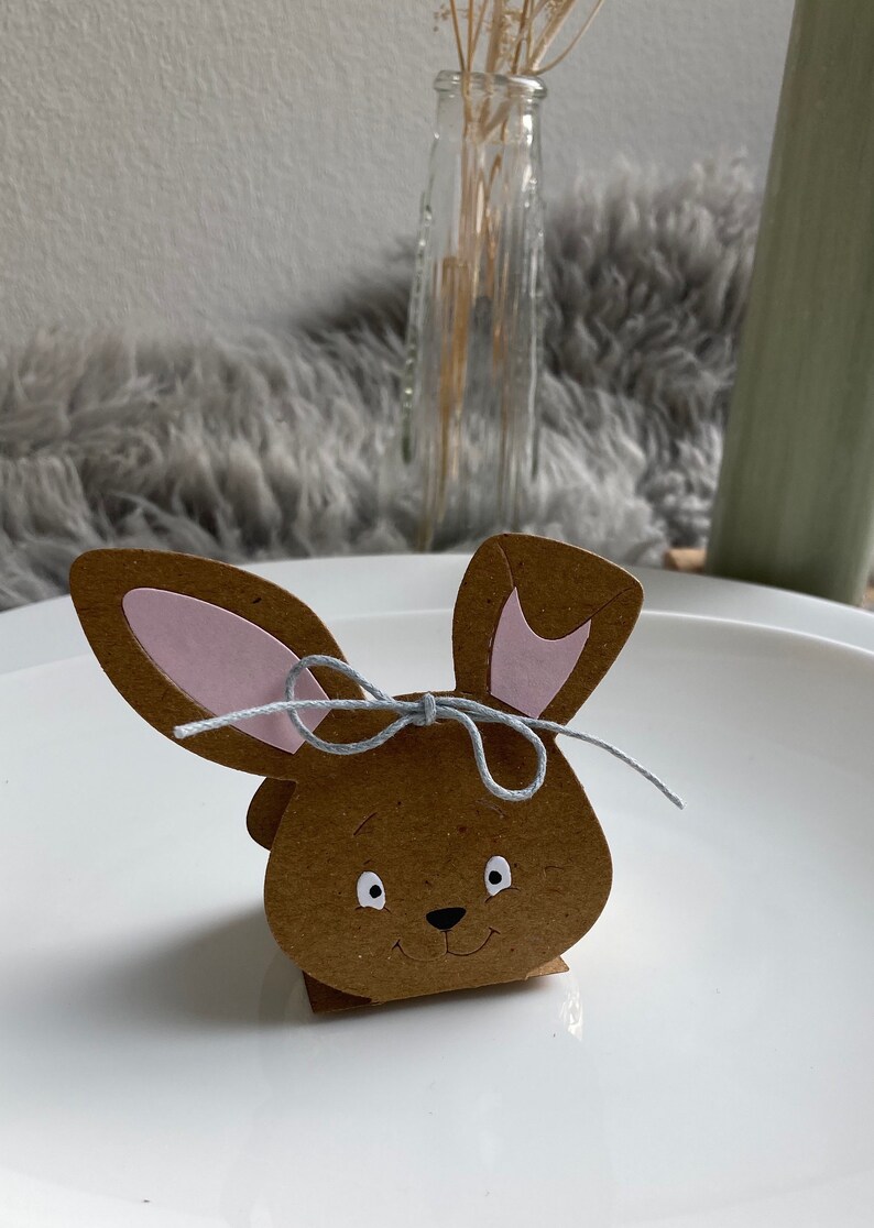 May include: A brown paper bunny-shaped box with pink ears and a gray ribbon. The box has a white face with black eyes and a nose.
