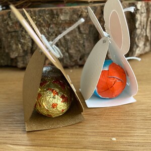 May include: Two small cardboard boxes shaped like bunnies, each holding a chocolate egg. One box is brown with a gold foil wrapped egg, the other is white with a blue and orange foil wrapped egg.