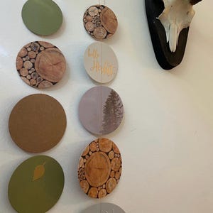 May include: A decorative garland featuring various circular ornaments. Some ornaments display wood grain patterns, while others are solid colors like green and brown. One ornament has the text "hello Herbst" in gold script. A deer skull is mounted on the wall.