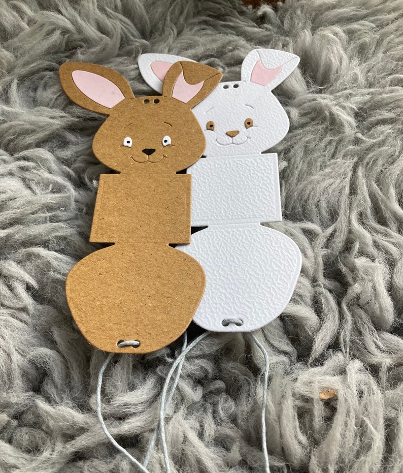 May include: Two bunny-shaped gift boxes, one brown and one white, with a string attached to each. The boxes are made of cardboard and have a textured surface.