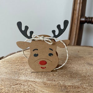 May include: A brown cardboard reindeer head with black antlers and a red nose. The reindeer is tied with a white twine bow.