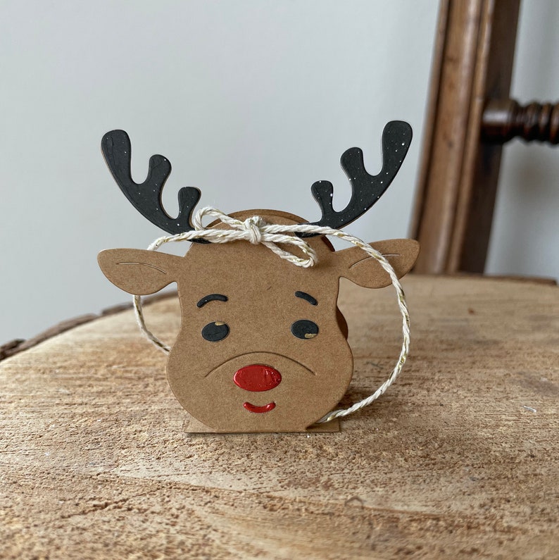 May include: A brown cardboard reindeer head with black antlers and a red nose. The reindeer is tied with a white twine bow.