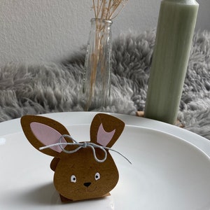 May include: A brown bunny-shaped cardboard box with pink ears and a gray ribbon tied around the neck. The box is sitting on a white plate.