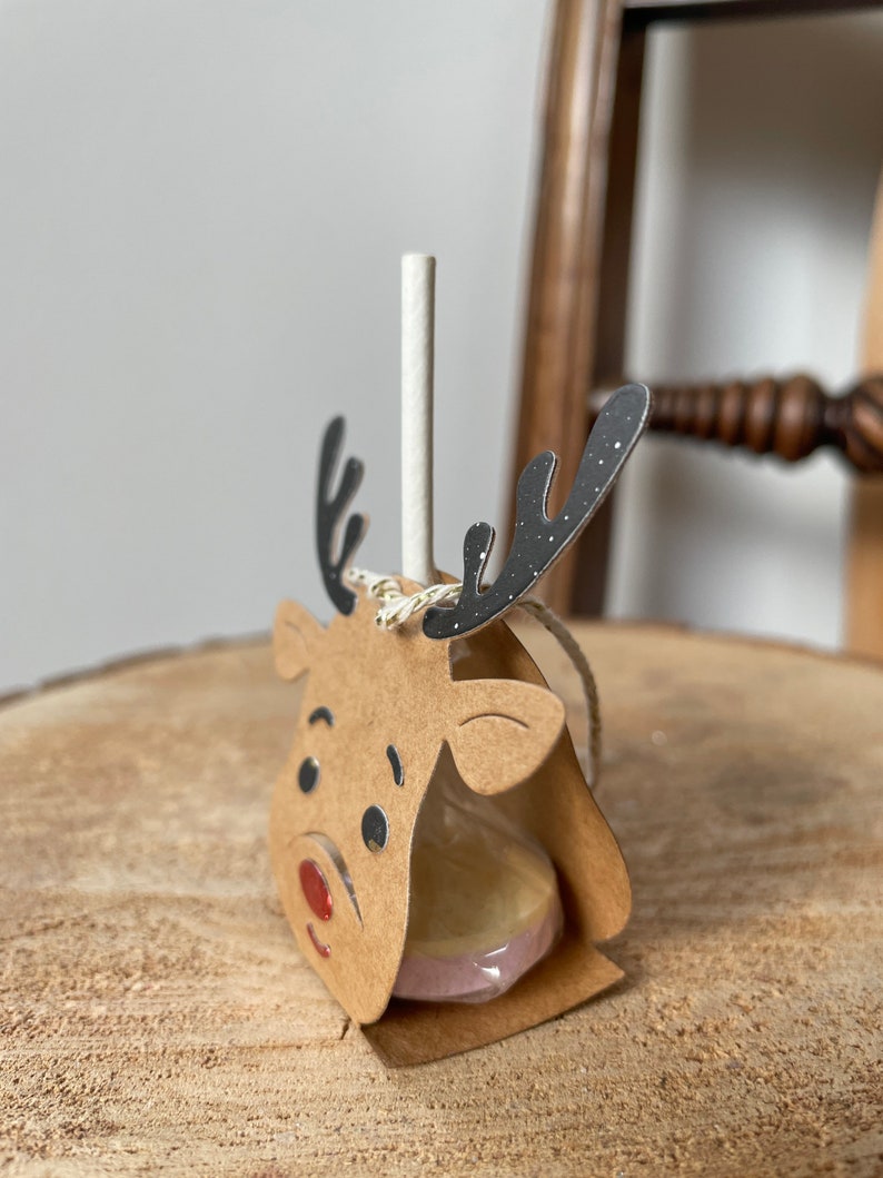 May include: A brown cardboard reindeer head with black antlers and a red nose holds a round lollipop. The lollipop is wrapped in clear plastic and has a white and pink swirl design.