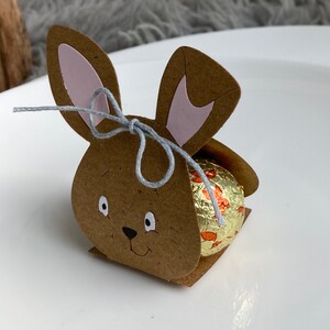 May include: A brown cardboard bunny rabbit box with pink ears and a gray string tied around the ears. The box is holding a chocolate egg wrapped in gold foil with orange and white designs.