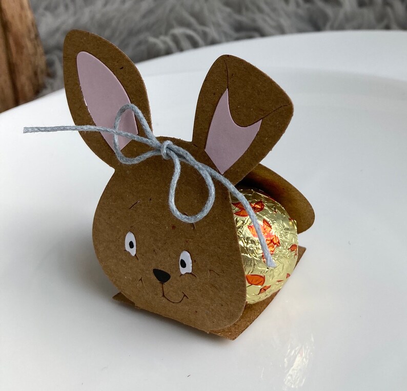 May include: A brown cardboard bunny rabbit box with pink ears and a gray string tied around the ears. The box is holding a chocolate egg wrapped in gold foil with orange and white designs.