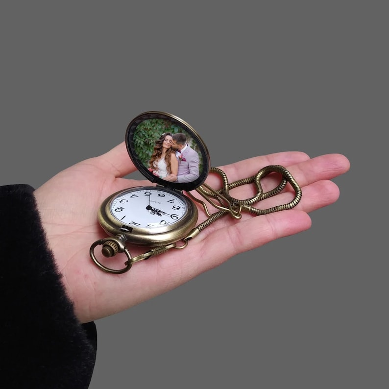 May include: Antique brass pocket watch with a white clock face and a photo inside the hinged cover. The watch is attached to a chain and is held open, revealing a picture of a couple. The watch has a classic, vintage design.