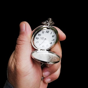May include: A silver pocket watch with a white face and black numerals is held open against a black background. The watch has a chain and a hinged cover. The watch face reads "QUARTZ".