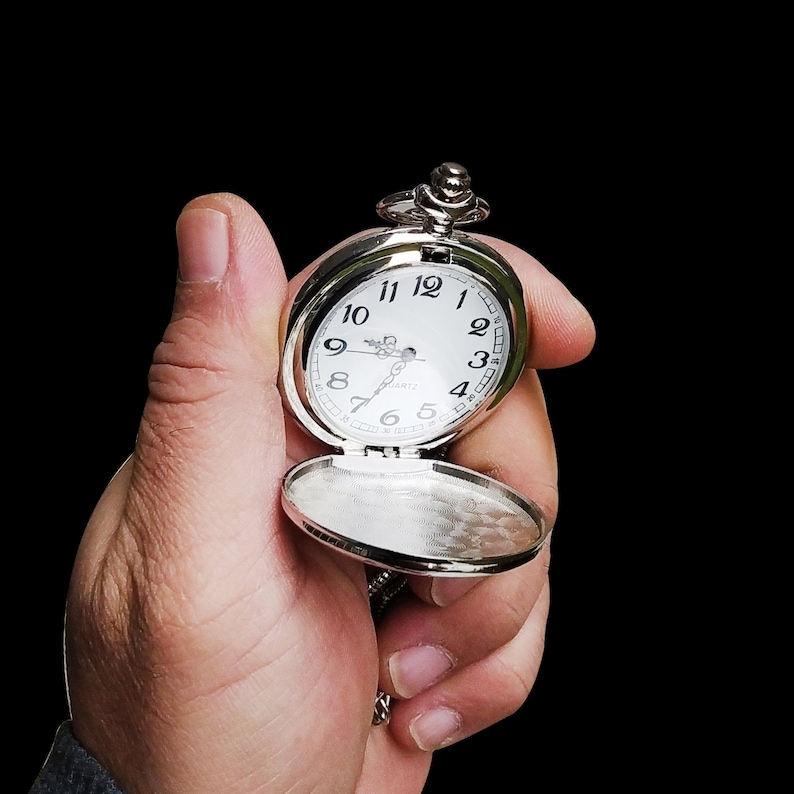 May include: A silver pocket watch with a white face and black numerals is held open against a black background. The watch has a chain and a hinged cover. The watch face reads "QUARTZ".