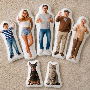 Op de afbeelding: Een verzameling op maat bedrukte lichaamskussens met afbeeldingen van mensen en huisdieren. De kussens tonen verschillende individuen in vrijetijdskleding, samen met een hond en een kat. De kussens zijn gerangschikt op een neutraal gekleurd tapijt.