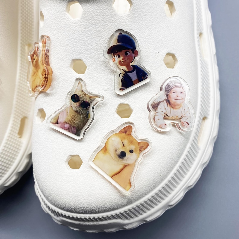 May include: White shoe with five colorful Jibbitz charms. The charms feature a cartoon boy, a cat wearing sunglasses, a dog with its tongue sticking out, and two babies.