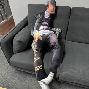 K&ouml;nnte beinhalten: Ein neuartiges Kissen in Form einer Person, die eine Jacke, ein gestreiftes Oberteil und eine dunkle Hose tr&auml;gt. Das Kissen liegt auf einem dunkelgrauen Sofa. Das Gesicht der Person ist auf dem Kissen zu sehen.